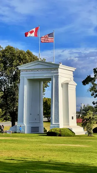 The Peace Arch stands on a green lawn beneath Canadian and United States flags, with the inscription “BRETHREN DWELLING TOGETHER IN UNITY” visible across its upper facade.