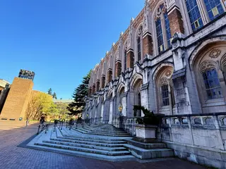 The Gothic stone-and-brick facade of Suzzallo Library rises above broad steps and a brick plaza beneath a clear blue sky.