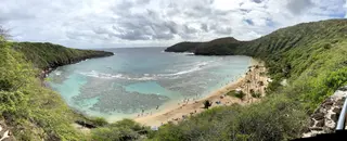 A sandy beach curves around a turquoise bay beneath lush green cliffs and a cloudy sky.