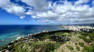 A panoramic view from a green hillside overlooks turquoise ocean water, a sandy coastline, and a dense cluster of high-rise buildings beneath dramatic clouds.