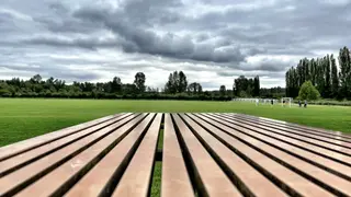 A wooden slatted bench overlooks a green sports field with distant players beneath a cloudy sky.