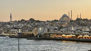 The Galata Bridge crosses choppy water beneath the illuminated Süleymaniye Mosque and Istanbul’s densely built skyline at dusk.