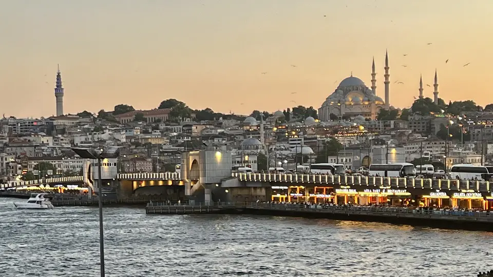 The Galata Bridge crosses choppy water beneath the illuminated Süleymaniye Mosque and Istanbul’s densely built skyline at dusk.