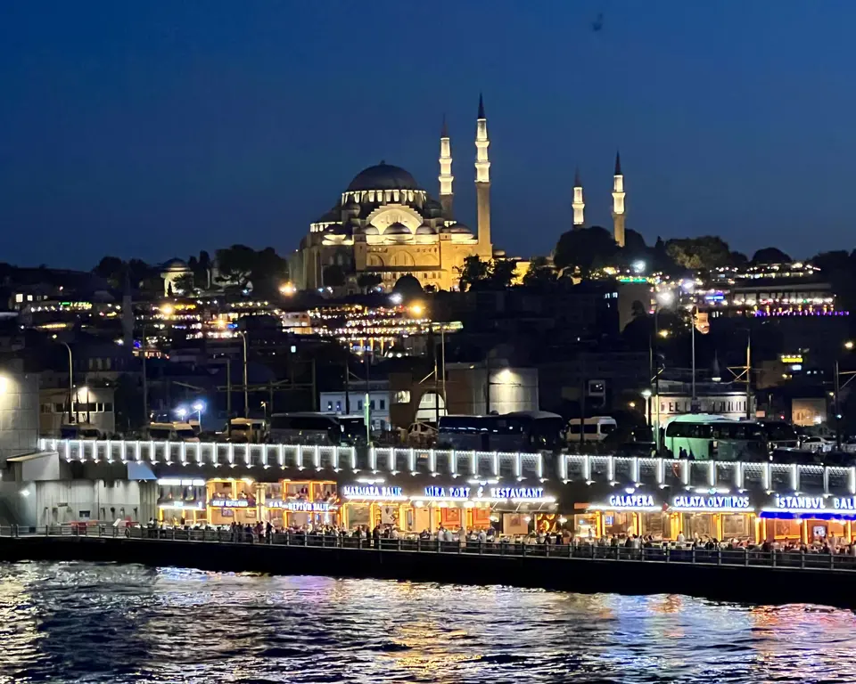 Hagia Sophia and its four illuminated minarets rise above a brightly lit waterfront of restaurants and rippling water at night.