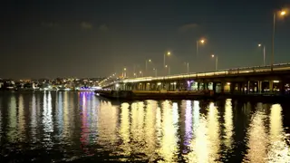 A long illuminated bridge spans dark water, casting bright reflections across the surface at night.