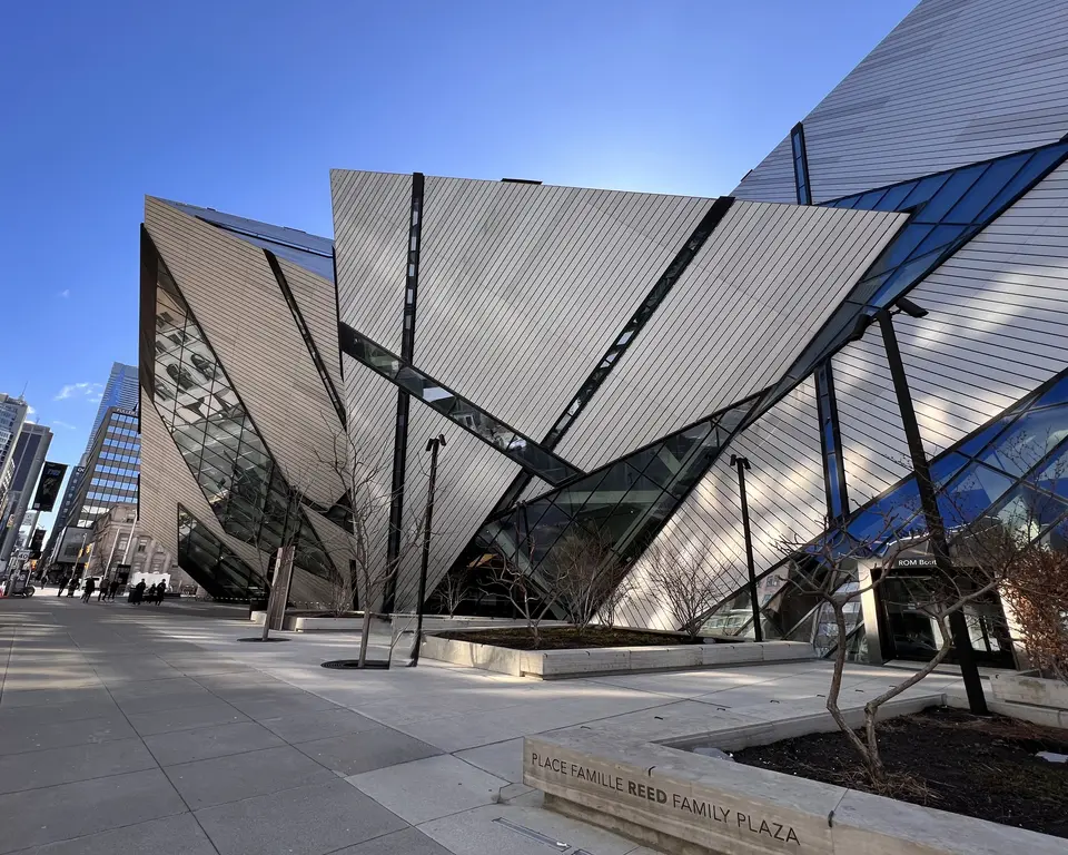 The angular crystalline facade of the Royal Ontario Museum rises beside the paved Place Famille Reed Family Plaza under a clear blue sky.