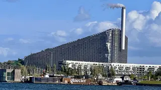 CopenHill rises behind waterfront buildings and boats, with a tall smokestack releasing white vapor beneath a blue sky.