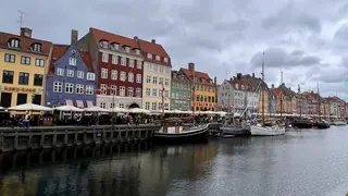 Colorful historic buildings, café terraces, and moored sailboats line the Nyhavn canal under a cloudy sky.