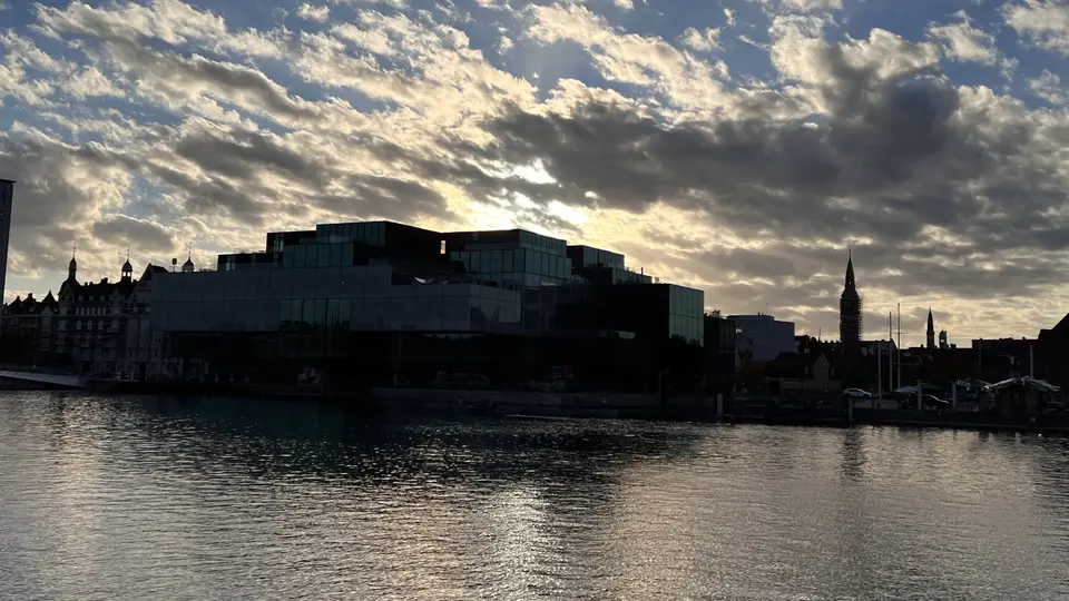 The Royal Danish Playhouse stands in silhouette along the waterfront beneath a dramatic cloud-filled sunset sky.