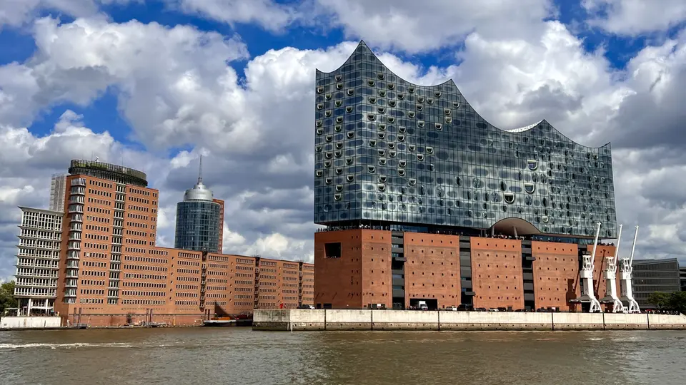 The Elbphilharmonie rises above red brick waterfront buildings beside the river under a dramatic cloudy sky.