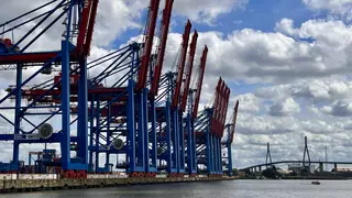 Container cranes labeled Terminal Burchardkai line the waterfront beneath a cloudy sky, with the Köhlbrand Bridge in the distance.