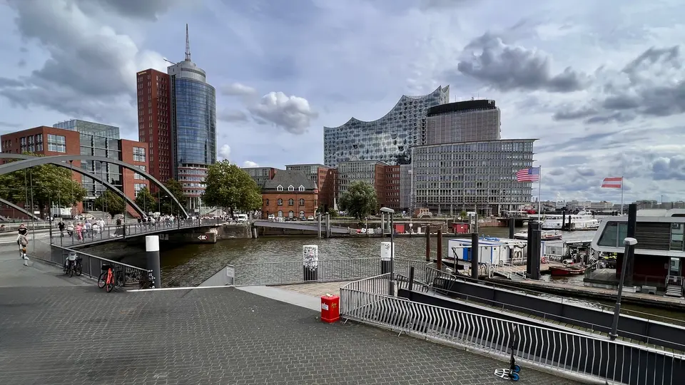 The Elbphilharmonie rises above a harbor basin with boats, a pedestrian bridge, and modern waterfront buildings beneath cloudy skies.