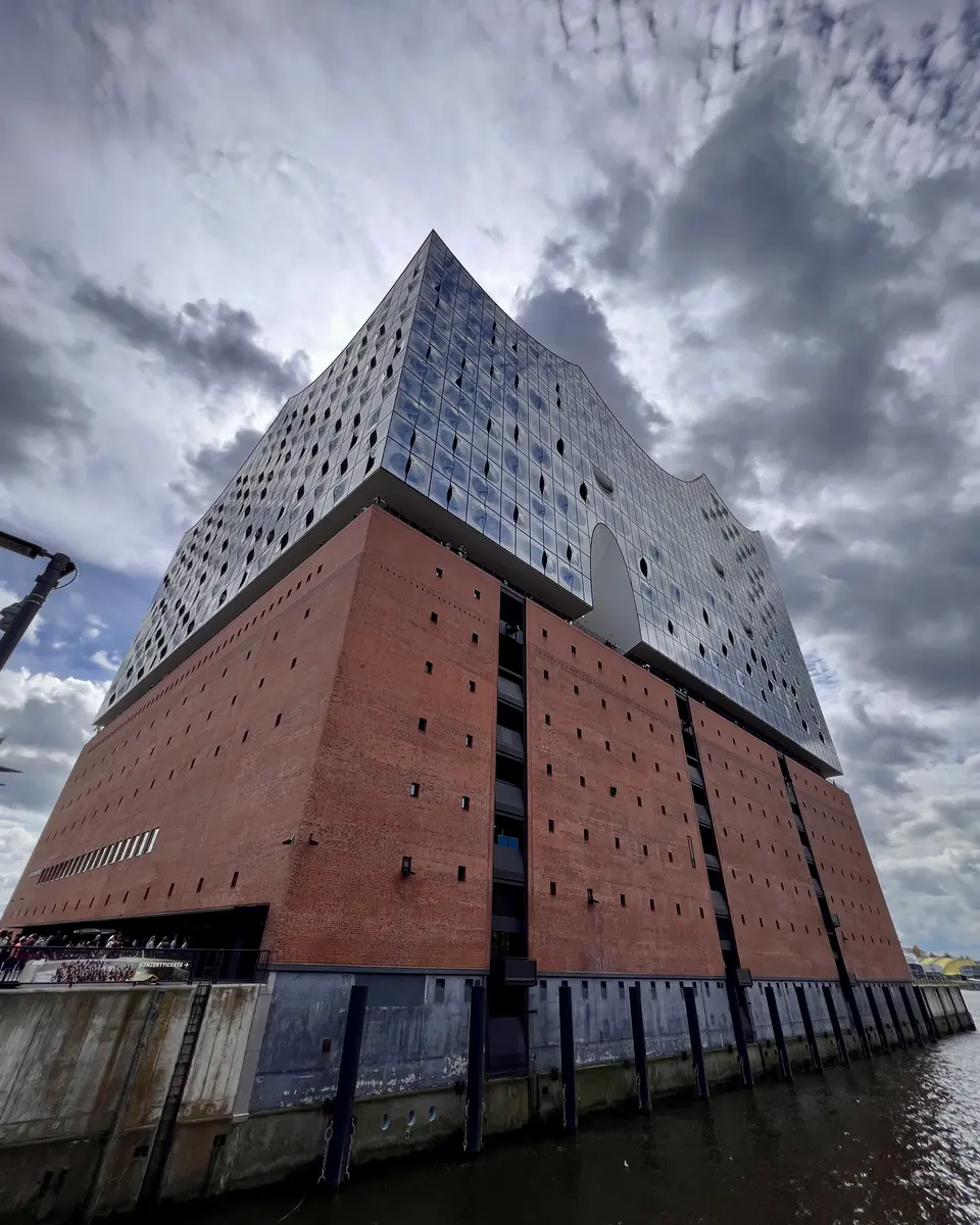 The Elbphilharmonie rises above the harbor water with its red brick base and rippling glass façade beneath dramatic clouds.