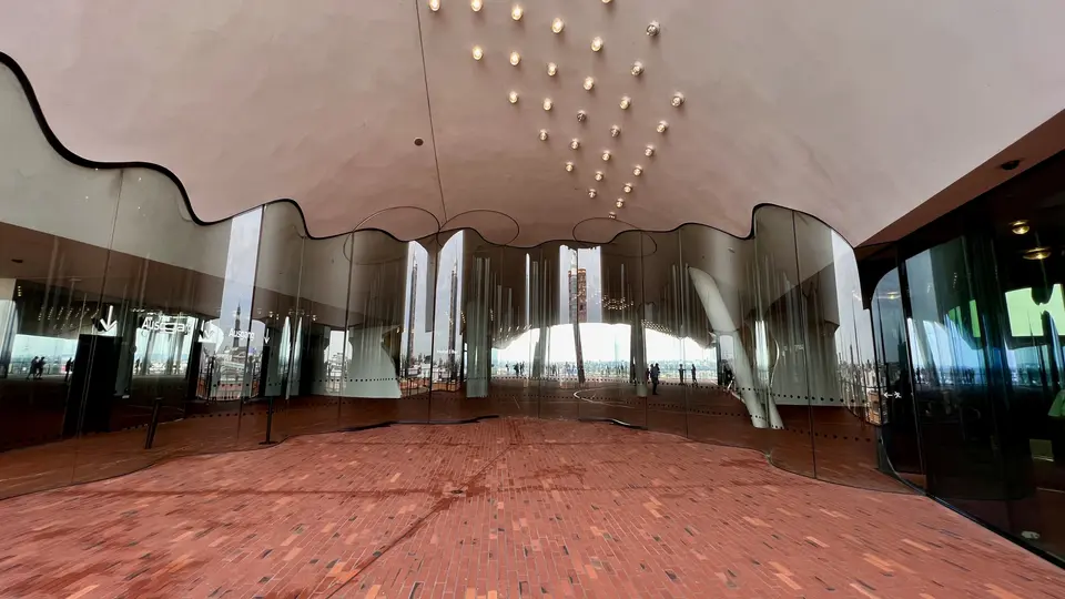 The Elbphilharmonie’s glass-walled plaza features a red brick floor, a wavy ceiling, and small hanging lights.