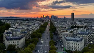 A tree-lined Paris boulevard leads toward a distant skyline beneath a dramatic orange sunset, with Tour Montparnasse rising on the right.