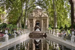 The Médici Fountain stands at the end of a reflecting pool beneath leafy trees, with flower-filled urns and visitors along the sides.
