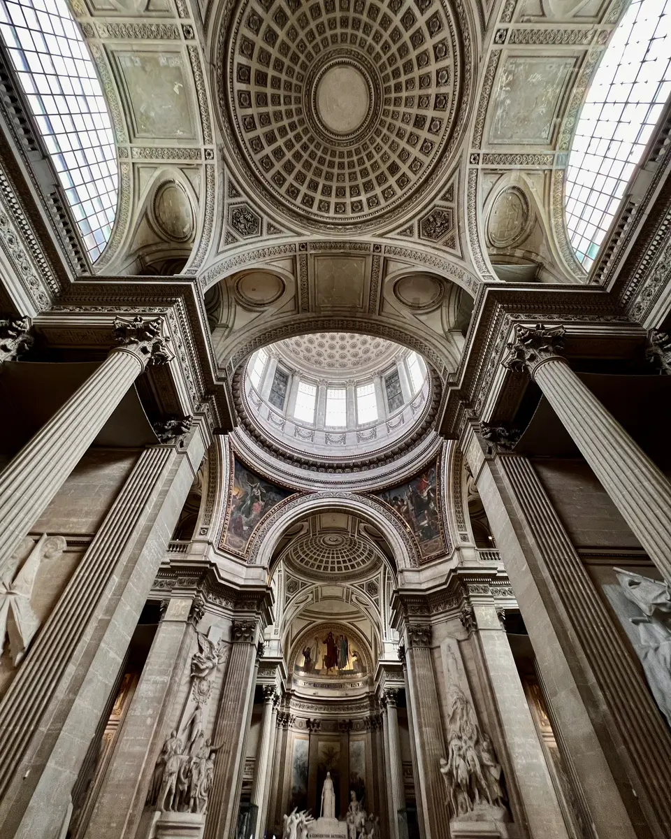 The ornate vaulted interior of the Panthéon features coffered domes, skylights, towering fluted columns, and sculptural reliefs.
