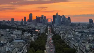 The Grande Arche de la Défense rises among the modern skyline beyond a tree-lined Paris avenue at sunset.