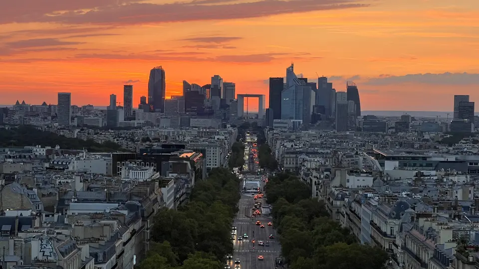 The Grande Arche de la Défense rises among the modern skyline beyond a tree-lined Paris avenue at sunset.