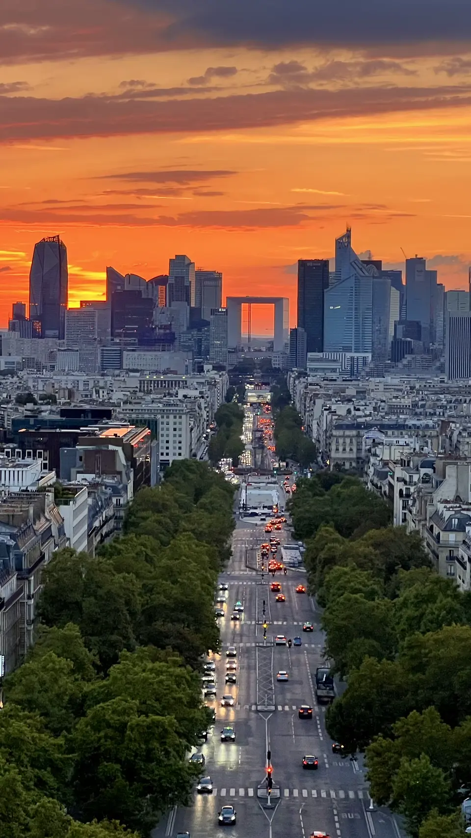 The Grande Arche de la Défense rises beyond a tree-lined boulevard filled with traffic beneath a vivid orange sunset.