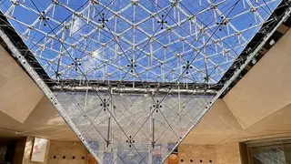 The inverted glass pyramid of the Pyramide Inversée du Louvre rises above a stone-lined interior beneath a blue sky.