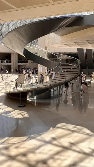 A sweeping spiral staircase curves through the sunlit Louvre Museum lobby beneath a glass ceiling as visitors gather below.
