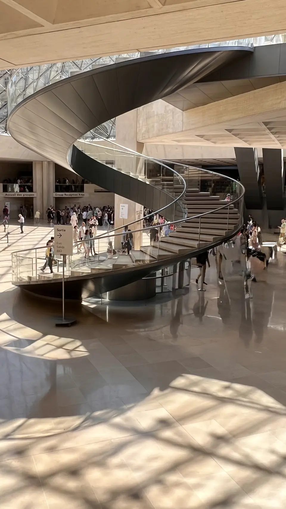 A sweeping spiral staircase curves through the sunlit Louvre Museum lobby beneath a glass ceiling as visitors gather below.