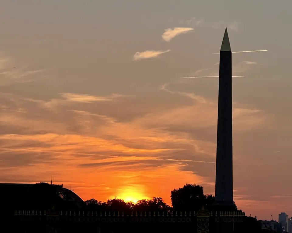 The Luxor Obelisk stands silhouetted against an orange sunset sky above dark treetops and rooftops.