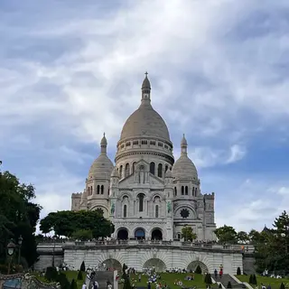 The white domes of the Sacré-Cœur Basilica rise above terraced gardens and visitors beneath a cloudy blue sky.