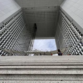 Stone steps lead up to the Grande Arche de la Défense, whose vast square opening frames a suspended canopy and scattered visitors.