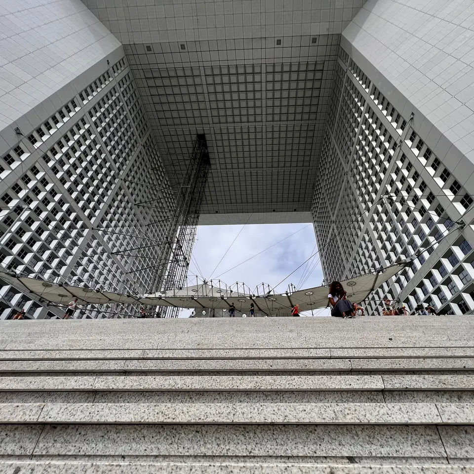 Stone steps lead up to the Grande Arche de la Défense, whose vast square opening frames a suspended canopy and scattered visitors.