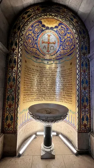 An ornate gold, blue, and red mosaic memorial panel inside the Sacré-Cœur Basilica frames a metal holy water font.