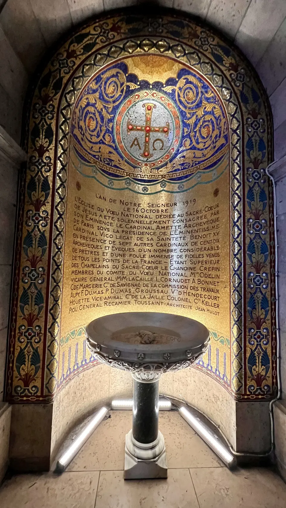 An ornate gold, blue, and red mosaic memorial panel inside the Sacré-Cœur Basilica frames a metal holy water font.