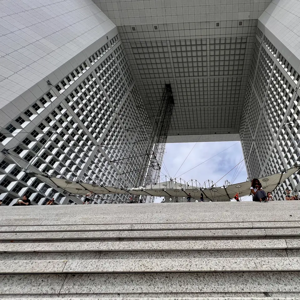 Broad stone steps rise beneath the towering rectangular arch of the Grande Arche de la Défense, with small pedestrians below its gridded facade.