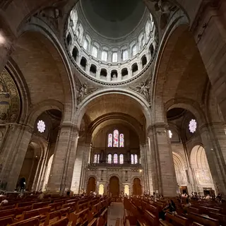 The Basilica of the Sacré-Cœur de Montmartre interior rises above rows of wooden pews with massive stone arches, stained-glass windows, and a lofty dome.