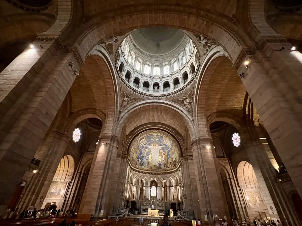 The interior of the Sacré-Cœur Basilica features soaring stone arches, a domed ceiling, and a large golden mosaic above the altar.