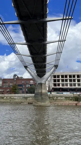 The underside of the Millennium Bridge stretches over the River Thames toward its central support, with riverside buildings beneath a cloudy blue sky.