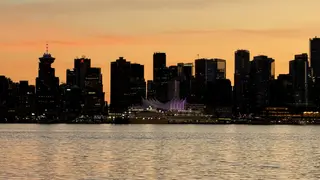 Vancouver's darkened skyline, including the sail-like roof of Canada Place, rises beyond calm water beneath an orange sunset.