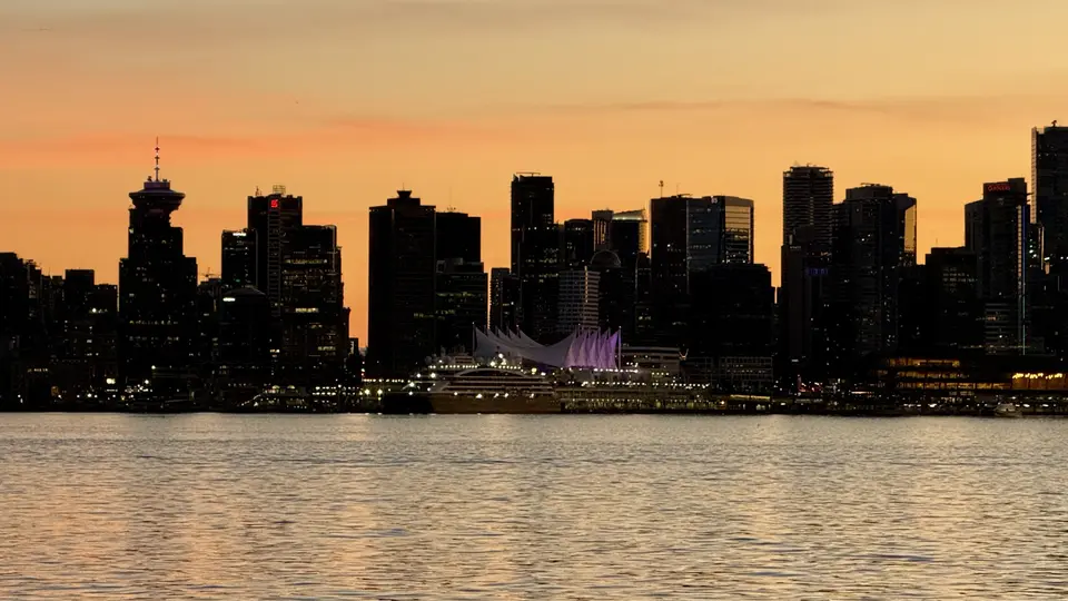 Vancouver's darkened skyline, including the sail-like roof of Canada Place, rises beyond calm water beneath an orange sunset.