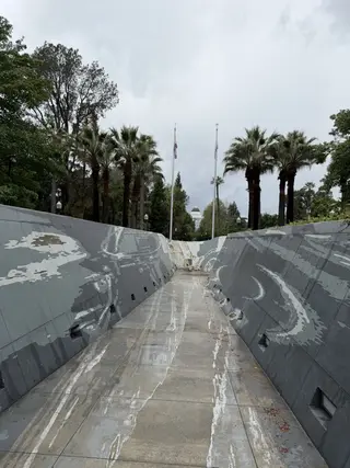 A rain-slicked memorial walkway with gray engraved walls leads through palm trees toward the California State Capitol dome.