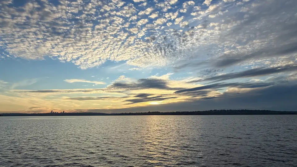 The setting sun glows beneath layered clouds over rippling water and a distant tree-lined shoreline.