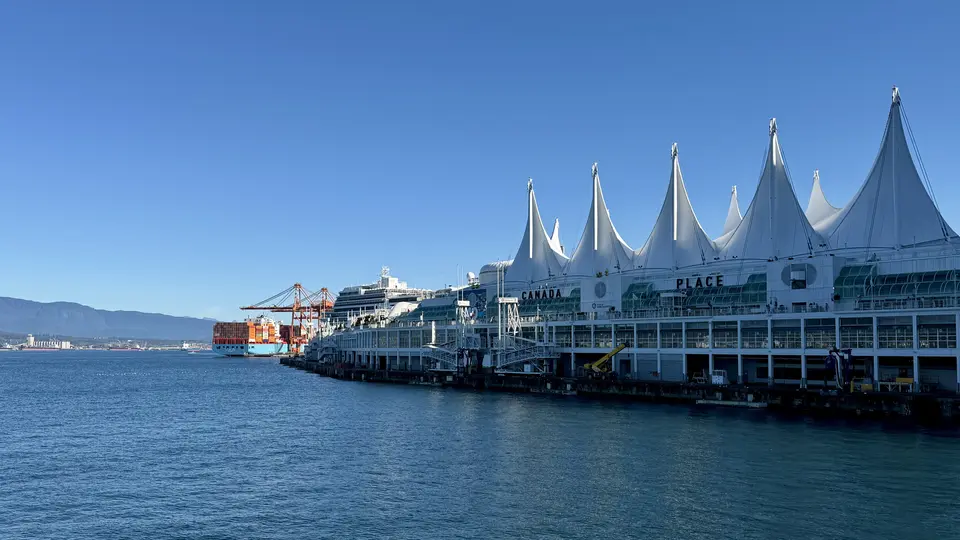 Canada Place’s distinctive white sails rise above the waterfront beside a cruise ship and cargo cranes under a clear blue sky.