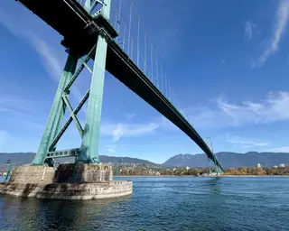 The Lions Gate Bridge spans blue water beneath a clear sky, with mountains and the distant shoreline behind it.