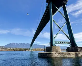 The Lions Gate Bridge spans blue water beneath a small airplane, with forested mountains in the distance.