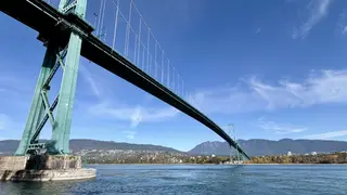 The Lions Gate Bridge spans blue water beneath a clear sky, with forested mountains and shoreline buildings in the distance.