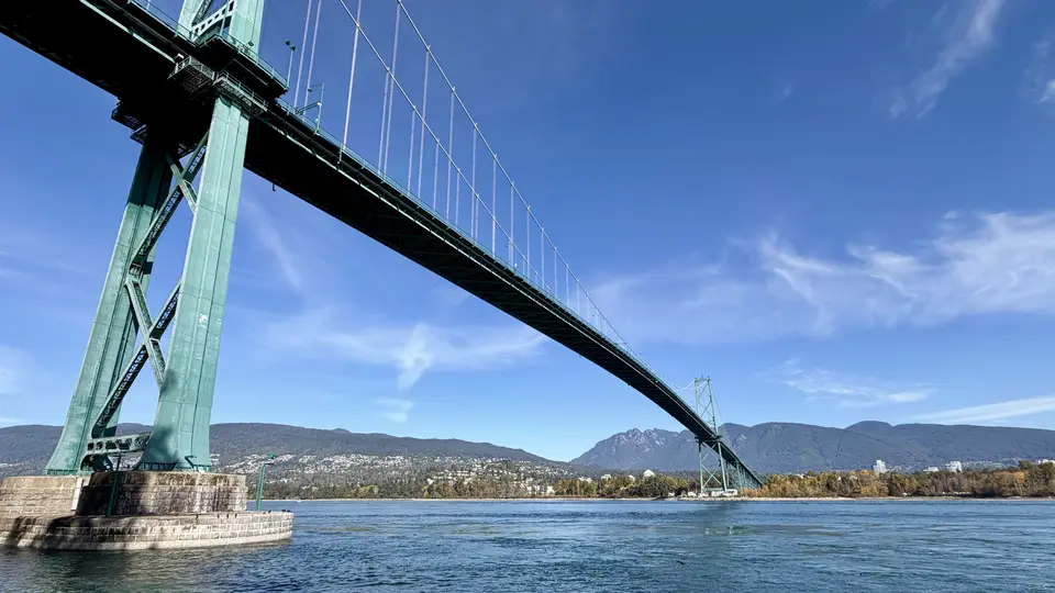 The Lions Gate Bridge spans blue water beneath a clear sky, with forested mountains and shoreline buildings in the distance.