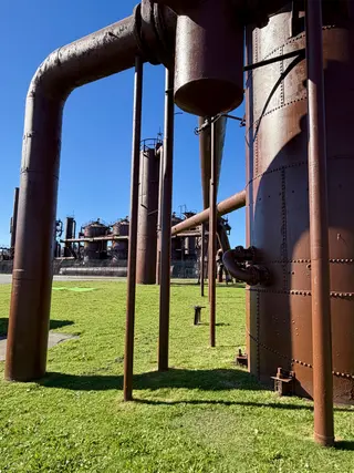 Rust-colored industrial pipes and riveted tanks stand above a grassy lawn beneath a clear blue sky.