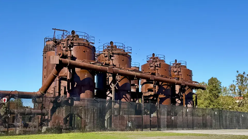 Rust-colored industrial machinery with cylindrical tanks and large pipes stands behind a black fence under a clear blue sky.